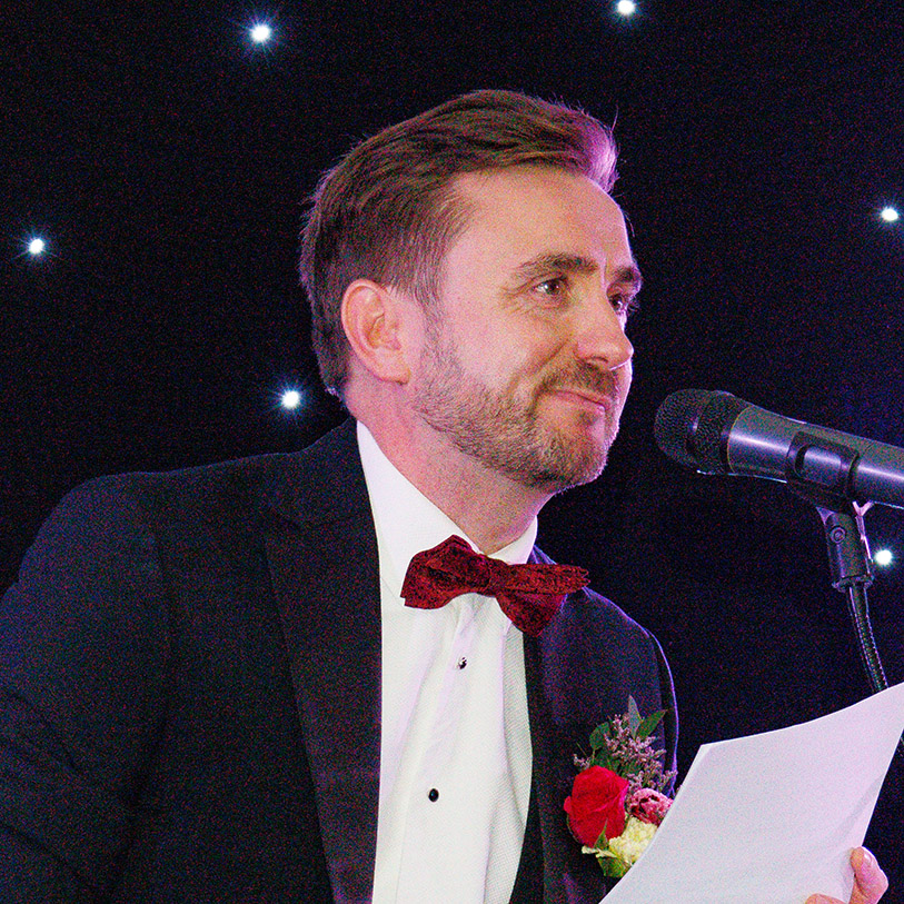 Kevin Durham in a tuxedo with a red bow tie speaking at a lectern microphone under a starlit backdrop Kevin Durham in a tuxedo with a red bow tie speaking at a lectern microphone under a starlit backdrop