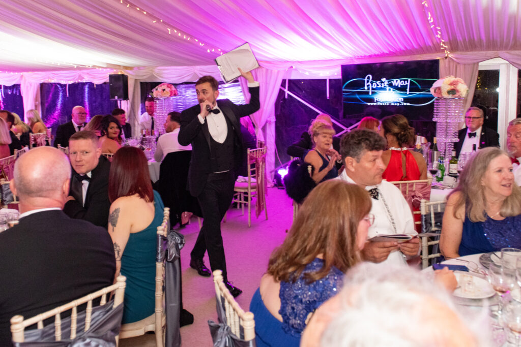 Charity auctioneer on the microphone holding up the lot sheet at a pink-lit marquee gala Charity auctioneer on the microphone holding up the lot sheet at a pink-lit marquee gala