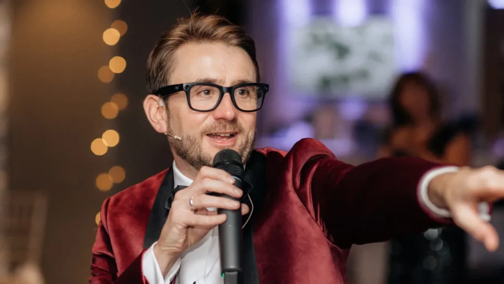 Kevin Durham in a red velvet dinner jacket, speaking into a microphone and gesturing to the room at a charity gala Kevin Durham in a red velvet dinner jacket, speaking into a microphone and gesturing to the room at a charity gala