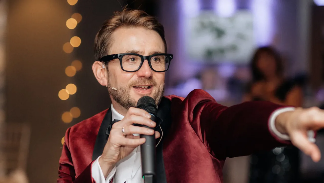 Kevin Durham in a red velvet dinner jacket, speaking into a microphone and gesturing to the room at a charity gala Kevin Durham in a red velvet dinner jacket, speaking into a microphone and gesturing to the room at a charity gala